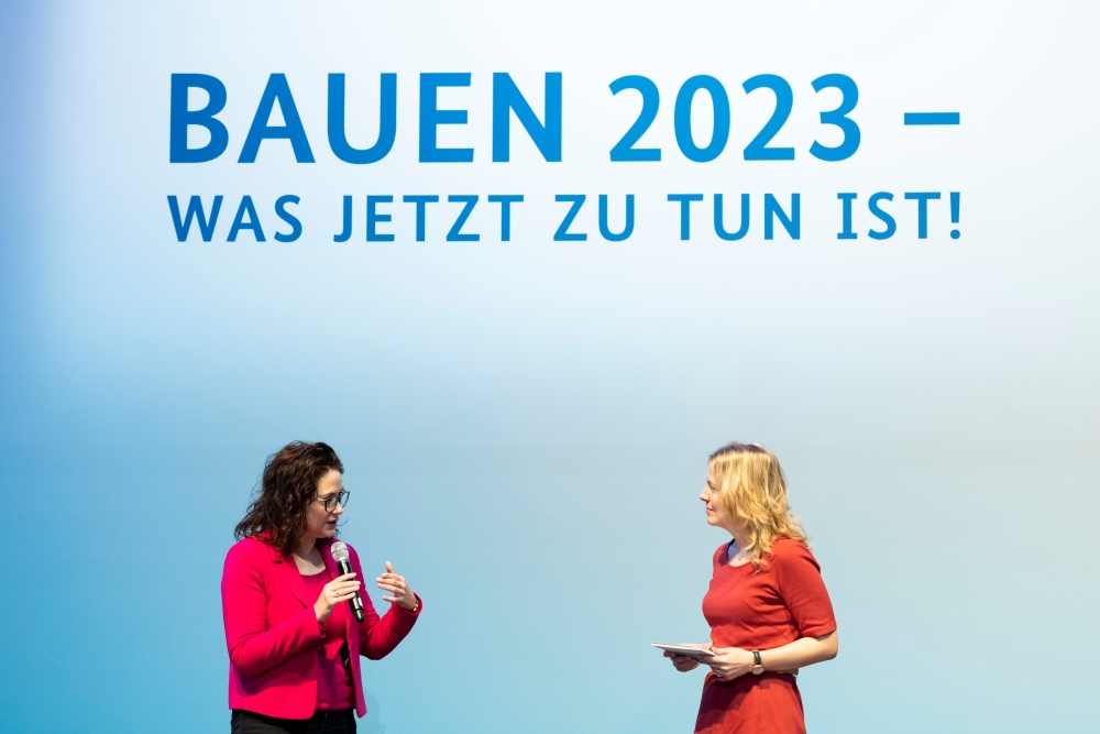 Tina Teucher spricht vor Publikum auf Messe BAU 2023; Text auf Hintergrund: BAUEN 2023 - Was jetzt zu tun ist!
